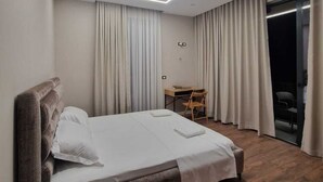 A modern hotel room with a large bed, a wooden nightstand, a desk, and a sliding glass door to a balcony. - Vila Kendezi (Vlorë)