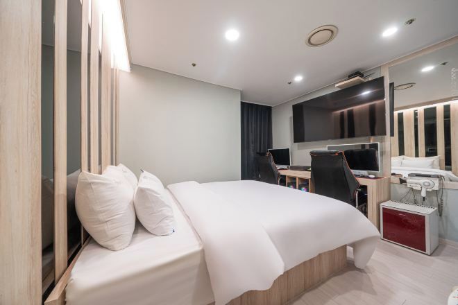 A modern hotel room with a bed, desk, chair, and a small desk with a computer and phone.