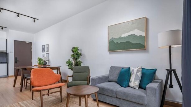 A modern living room with a grey sofa, orange chair, and a round wooden table. A painting of mountains hangs on the wall, and a lamp is on a tripod stand.