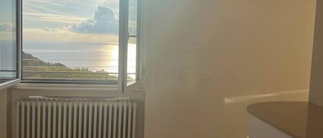 Una habitación con un amplio ventanal que da a una masa de agua y un paisaje distante. Hay un radiador blanco debajo de la ventana y un reloj en la pared.