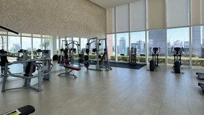 A spacious gym with various exercise equipment, large windows offering a view of the city, and a high ceiling. - Ritz Residence Platinum KL (Kuala Lumpur)