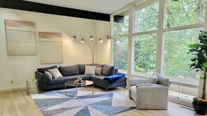 Un salon moderne avec un canapé foncé, une table basse et une grande fenêtre donnant sur un paysage verdoyant.