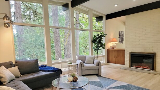 Un salon moderne comprenant un canapé, un fauteuil et une table basse, de grandes fenêtres offrant une vue sur les arbres, une cheminée et une lampe décorative.