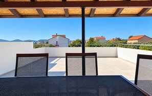 Un patio exterior con mesa y sillas, una pérgola arriba y vistas a casas y montañas al fondo.