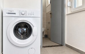 Una lavadora de carga frontal en un cuarto de lavado, con un inodoro y una ducha a la vista al fondo.