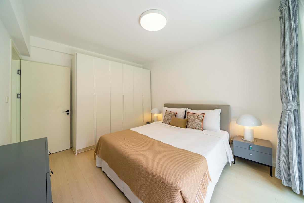 A bedroom with a large bed, a bedside table with a lamp, a wardrobe, and a curtain.
