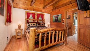 A wooden cabin bedroom with a bed, a nightstand, a TV mounted on the wall, and a painting of a bear.