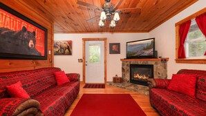 A cabin interior with two red patterned sofas, a stone fireplace, a ceiling fan, and a large bear painting on the wall.