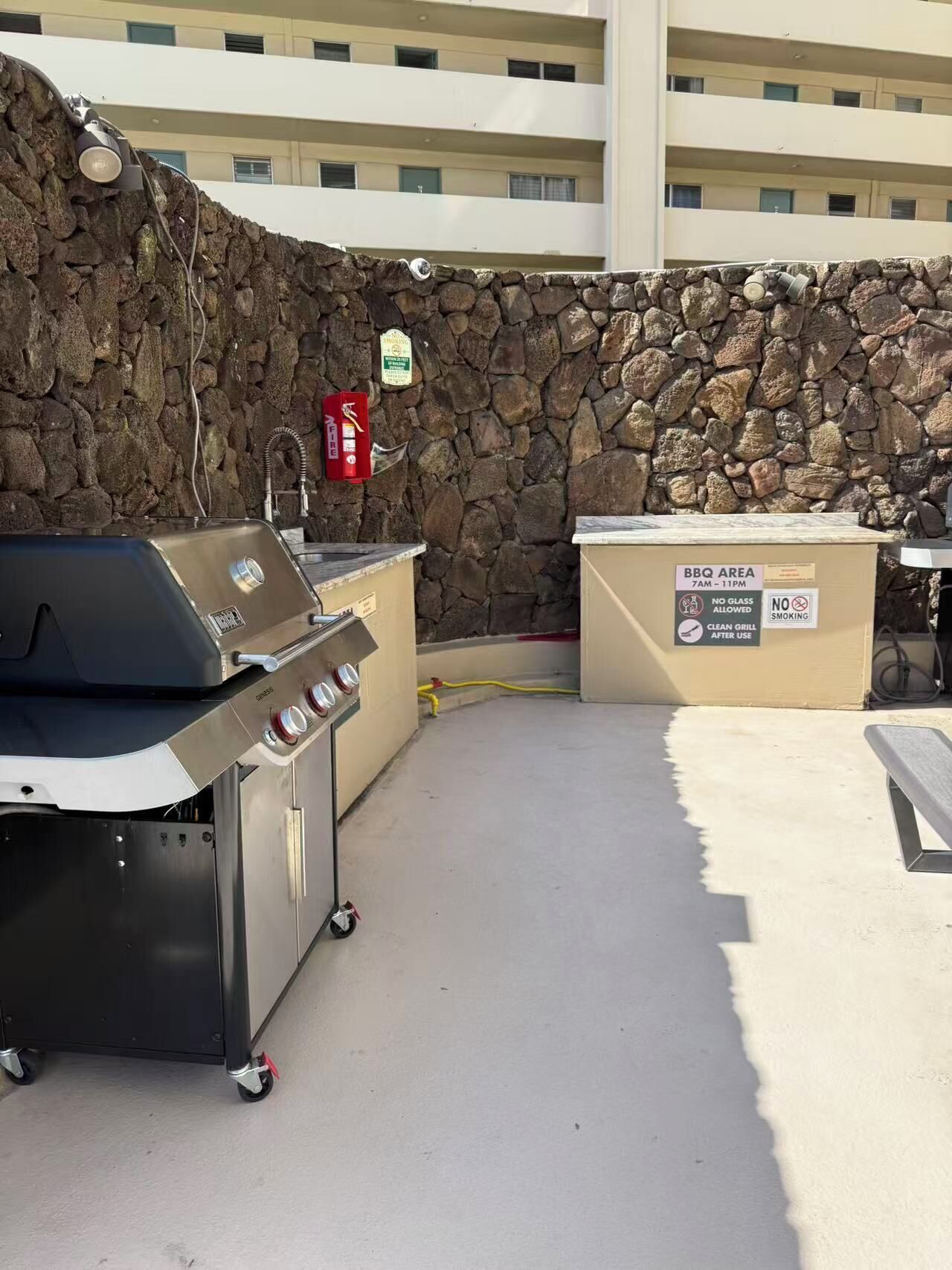 Een terras buiten met een barbecueplaats, een steenmuur, een grill en een bord met de barbecue tijden en regels.