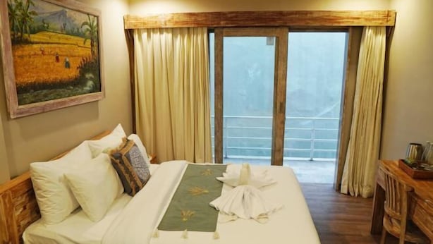 A hotel room with a bed, a painting on the wall, a balcony with curtains, and a wooden desk. - Koura Jimbaran (Bali)