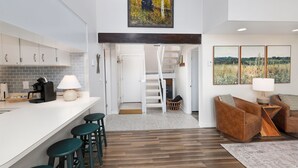Eine moderne Küche mit weißer Arbeitsplatte, Barhocker und einer Kaffeemaschine. Eine Treppe führt zu einer Erkerwohnung mit Blick ins Freie.
