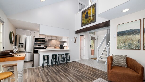 Eine moderne Küche mit Edelstahlgeräten, ein Essbereich mit Hockern und ein Wohnzimmer mit braunem Sofa und Teppich.