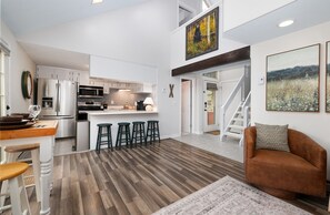 Una cocina moderna con electrodomésticos de acero inoxidable, un comedor con taburetes y una sala de estar con un sofá marrón y una alfombra.