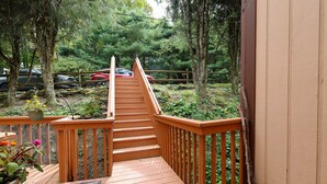 Eine Holzterrasse mit einer Treppe zu einem überdachten Bereich, umgeben von Bäumen und einem Parkplatz mit Autos.