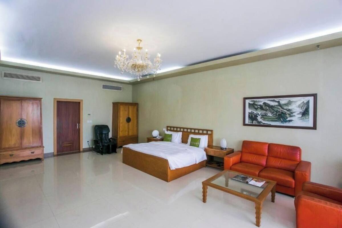 A hotel room with a large bed, a red leather sofa, a wooden coffee table, a chandelier, and a painting on the wall.