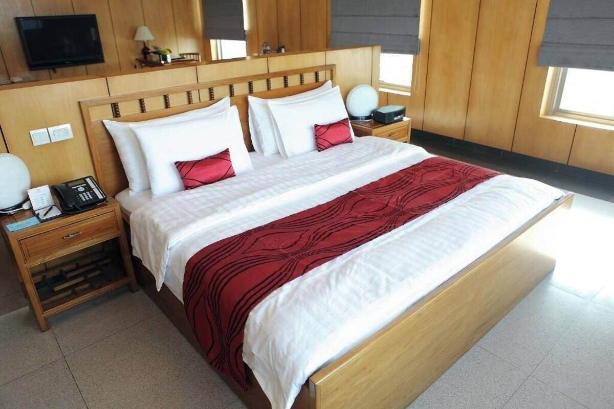 A hotel room with a large bed, wooden headboard, bedside tables with lamps, a TV, and a window with blinds.