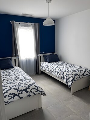 Une chambre avec deux lits simples, une fenêtre avec des rideaux, un luminaire au plafond et un mur bleu.