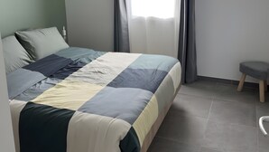 Une chambre à coucher avec un lit, un petit tabouret, une fenêtre avec des rideaux et un luminaire suspendu.