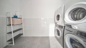 A laundry room with two stacked front-loading washing machines, a white tiled wall, and a metal shelf with a basket. - Express by M Village Tran Quoc Thao, a brand of Modern Village Lifestyle (Ho Chi Minh City)
