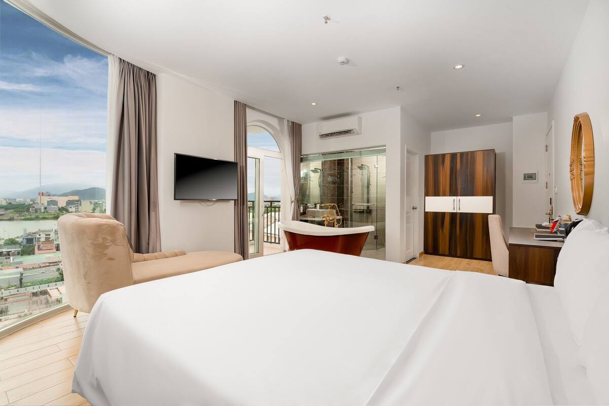 A modern hotel room with a large bed, a sitting area, a bathroom with a glass shower, and a city view.