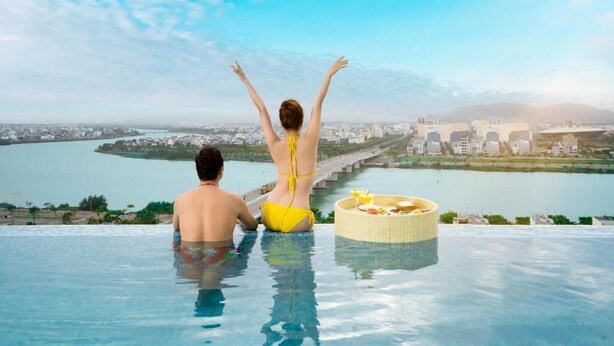 A couple in an infinity pool with a city view, a wicker basket with food, and a clear sky. - RHM Luxury Hotel and Suite  (Da Nang)