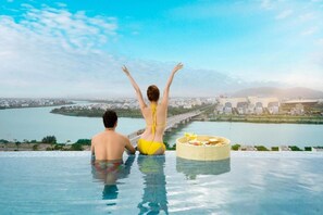 A couple in an infinity pool with a city view, a wicker basket with food, and a clear sky. - RHM Luxury Hotel and Suite  (Da Nang)