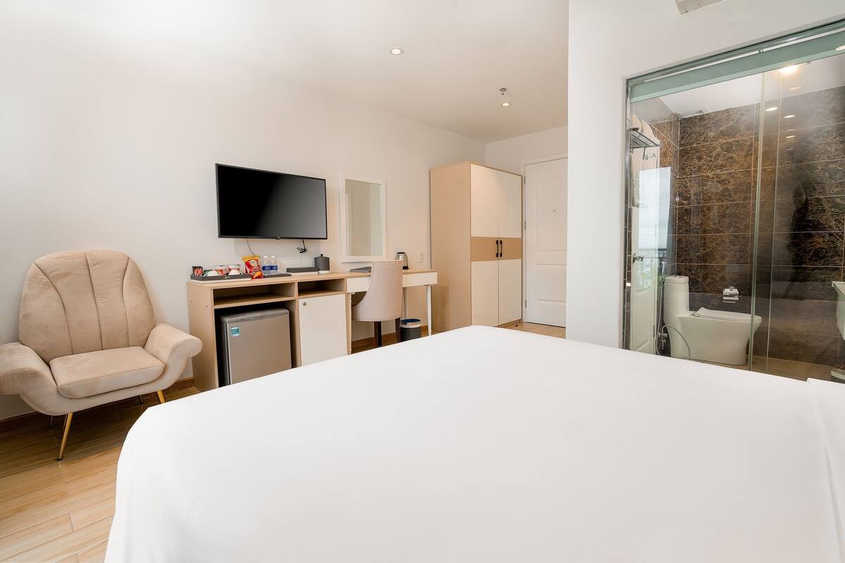 A modern hotel room with a bed, a TV, a desk with a computer, a chair, and a bathroom with a glass shower.