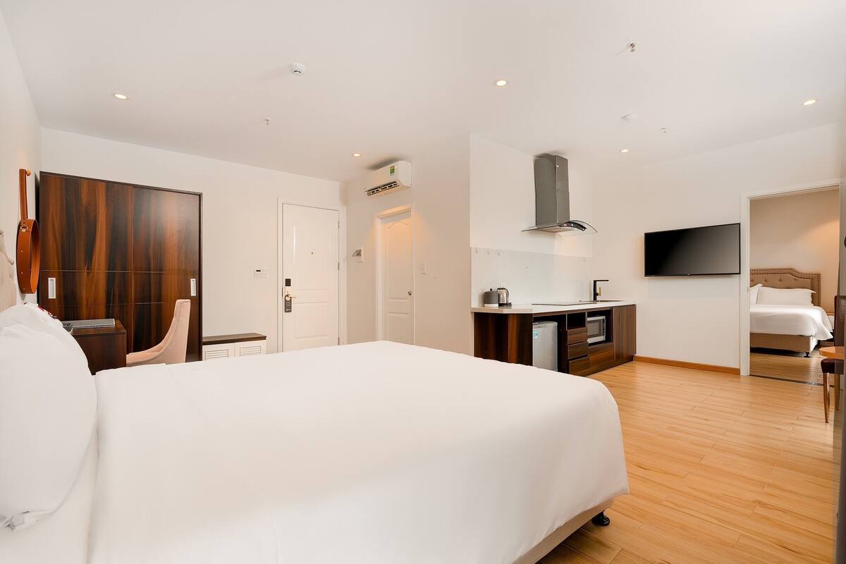 A modern hotel room with a large bed, a wooden wardrobe, a kitchenette, and a TV mounted on the wall.