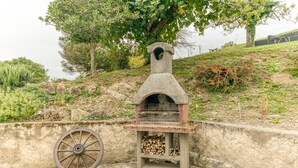 Un barbecue extérieur en pierre, doté de roulettes, avec des bûches et un arbre en arrière-plan.