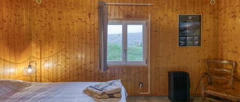 Une pièce aux murs lambrissés, avec un lit, un radiateur, une chaise, une fenêtre donnant sur un paysage et une affiche accrochée au mur.