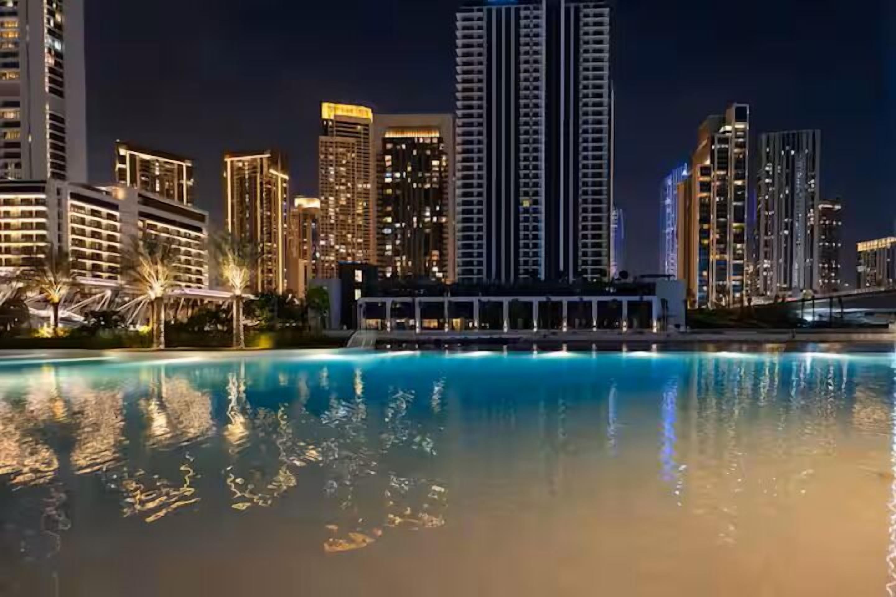 Nachts beleuchtete Stadtansicht mit Hochhäusern und einem Pool, der die Lichter widerspiegelt.