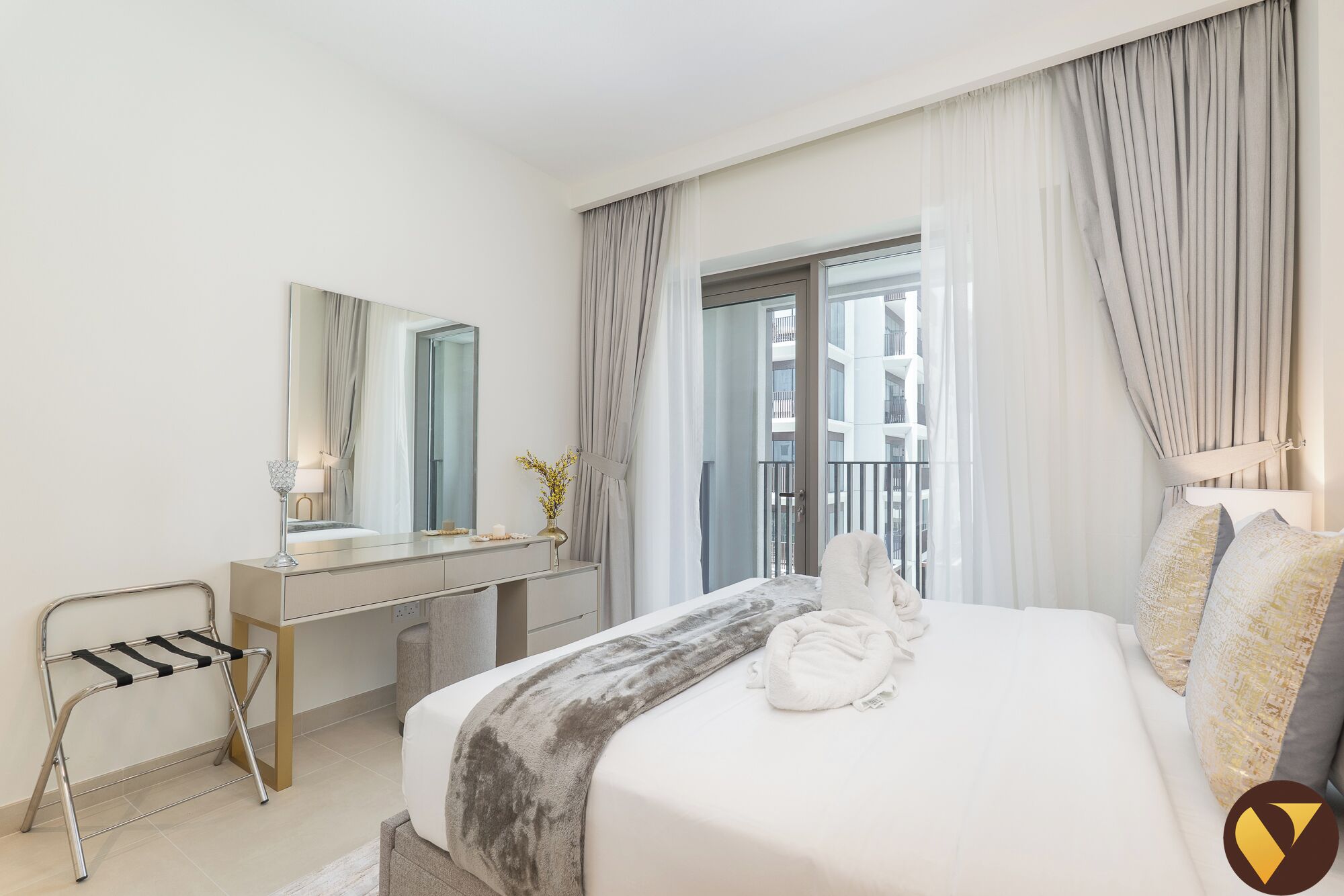 A hotel room with a bed, a vanity with a mirror, a window with curtains, and a view of the building outside.