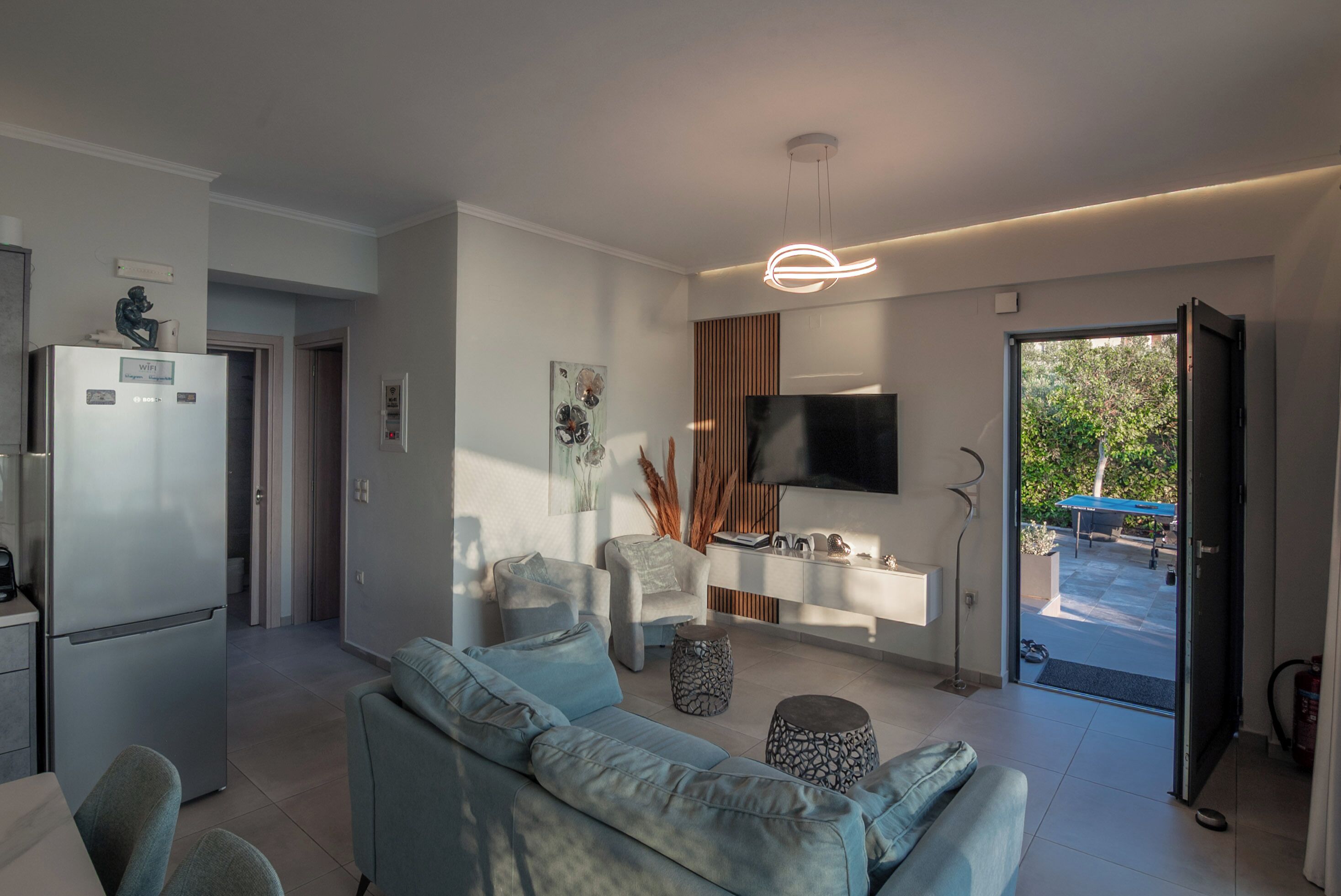 A modern living room with a sofa, a coffee table, a TV mounted on the wall, and a kitchen area with a refrigerator.