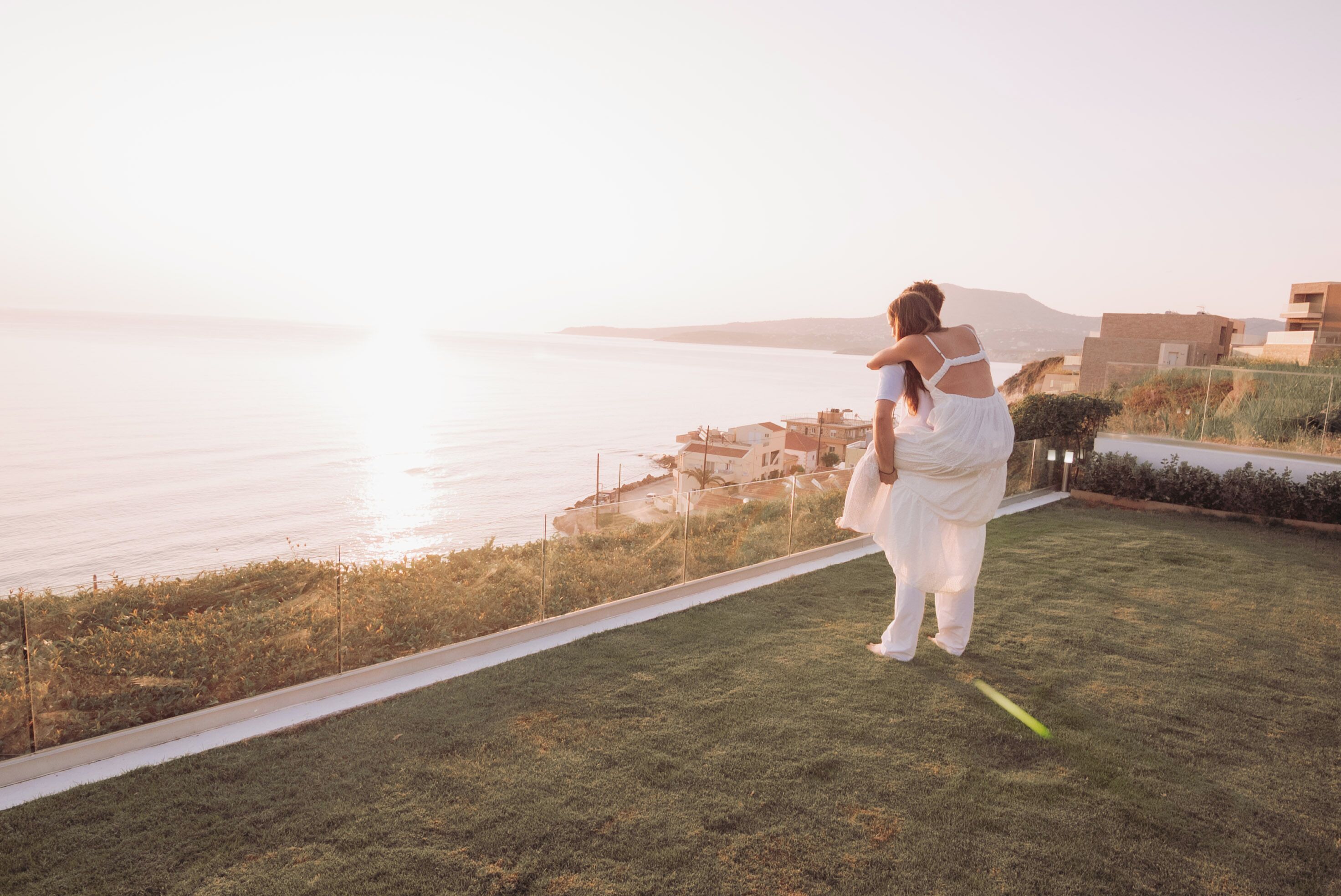 A person in a white dress standing on a grassy area with a view of the sea and buildings in the background.