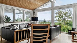 Ein modernes Wohnzimmer mit Holzboden, großen Fenstern mit Blick auf Grünflächen und Wasser, einem schwarzen Ledersofa und einem Holzstuhl.