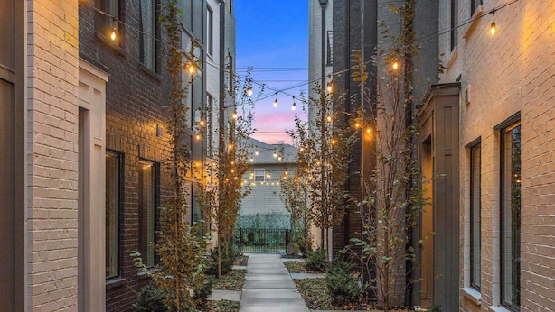 Une ruelle étroite bordée d’immeubles en briques, illuminée de guirlandes lumineuses, et un sentier menant à un portail.