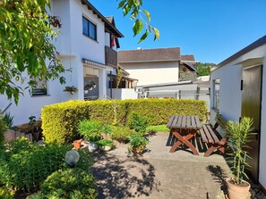 Une cour avec une table en bois et des bancs, entourée de plantes en pot et d’un arbuste taillé avec soin.