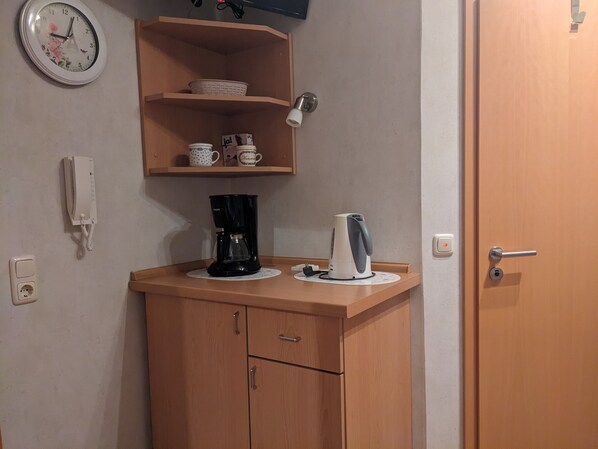 Une petite cuisine équipée d’une machine à café, d’une bouilloire et d  ’une étagère avec des tasses et un panier.