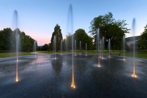 Una fontana con numerosi getti d'acqua disposti a cerchio, circondata da una zona pavimentata con panchine e alberi.