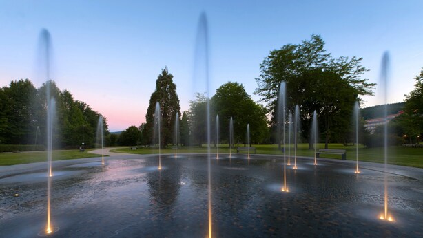 A fountain with multiple water jets in a circular pattern, surrounded by a paved area with benches and trees.