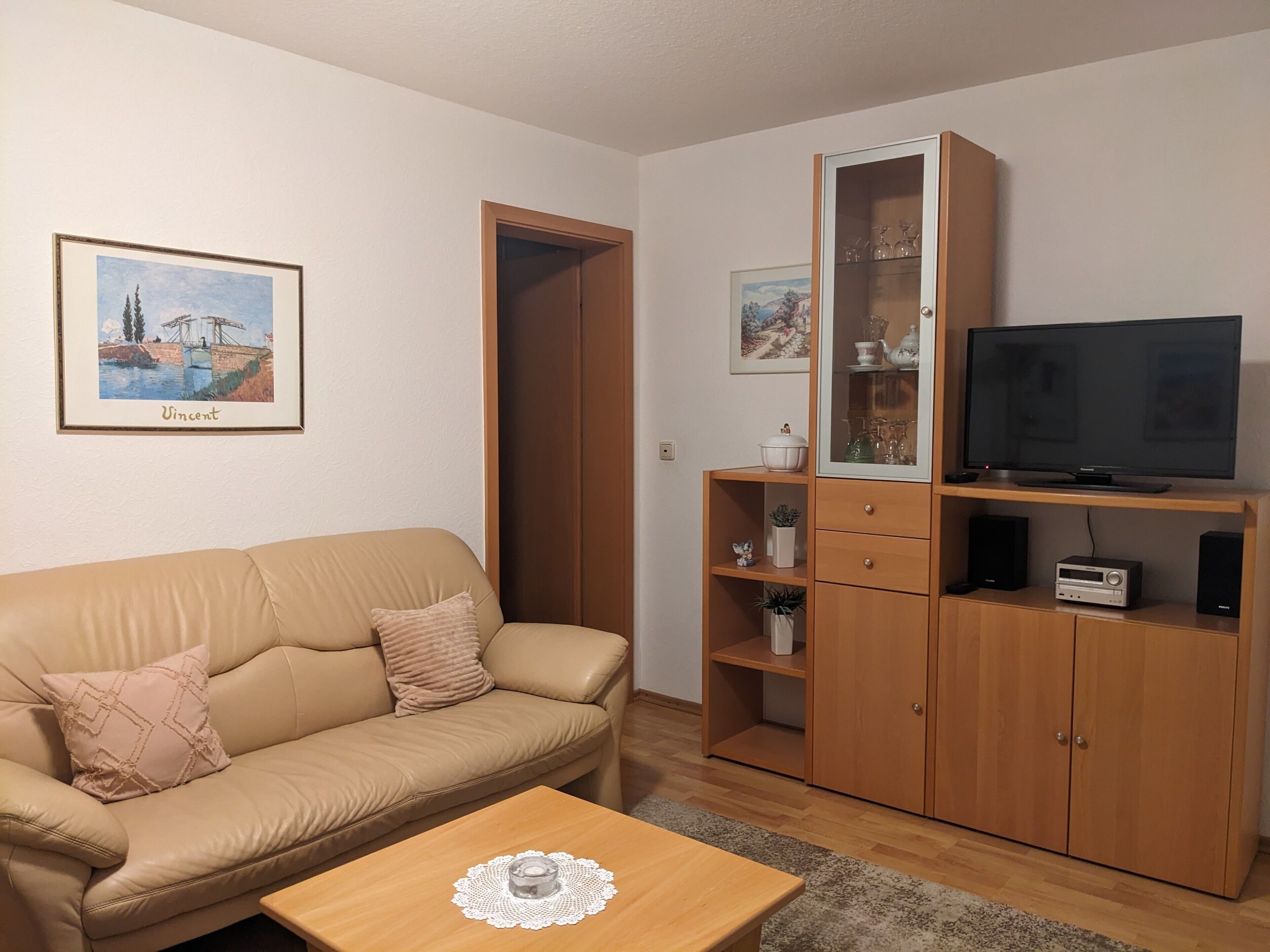 Ett vardagsrum med en beige soffa, ett träskåp med glasdörrar, en tv och inramade konstverk på väggen.