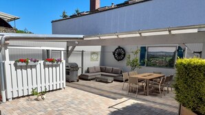 A covered patio with a white fence, outdoor seating, a table, and a view of the countryside.