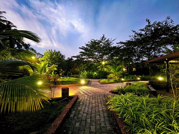 A paved pathway leading through a lush garden with various plants and trees, surrounded by a rustic building structure. - Arthigamya Spa & Resort (Gokarna)