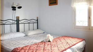 Un dormitorio con cama, ropa de cama blanca, colcha roja, un cuadro en la pared y una ventana con cortinas.
