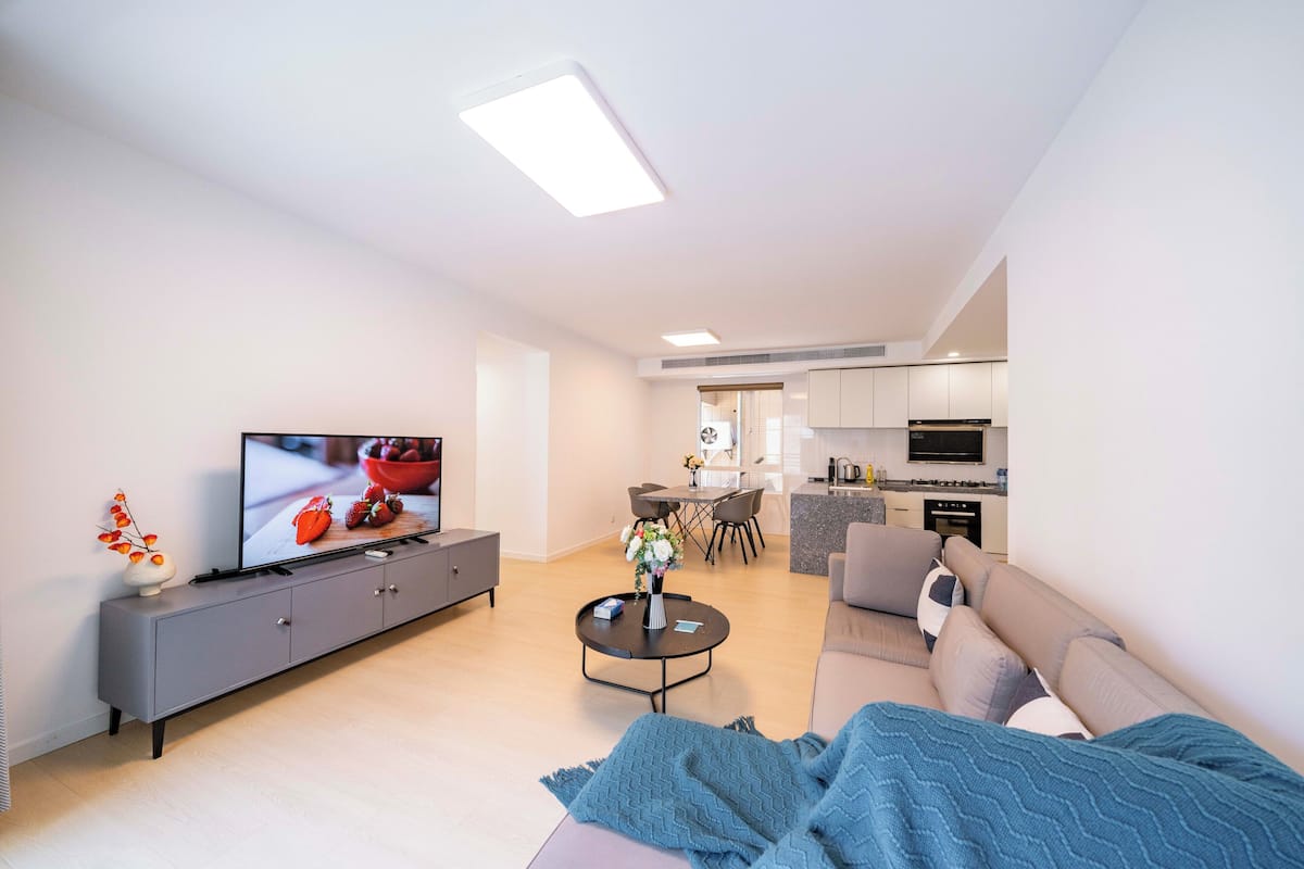 A modern living room with a flat-screen TV, grey sofa, and a dining area in the background.