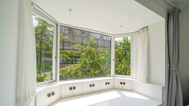 Een moderne kamer met een groot erkerraam, ingebouwde opbergruimte en uitzicht op een boom en gebouwen.