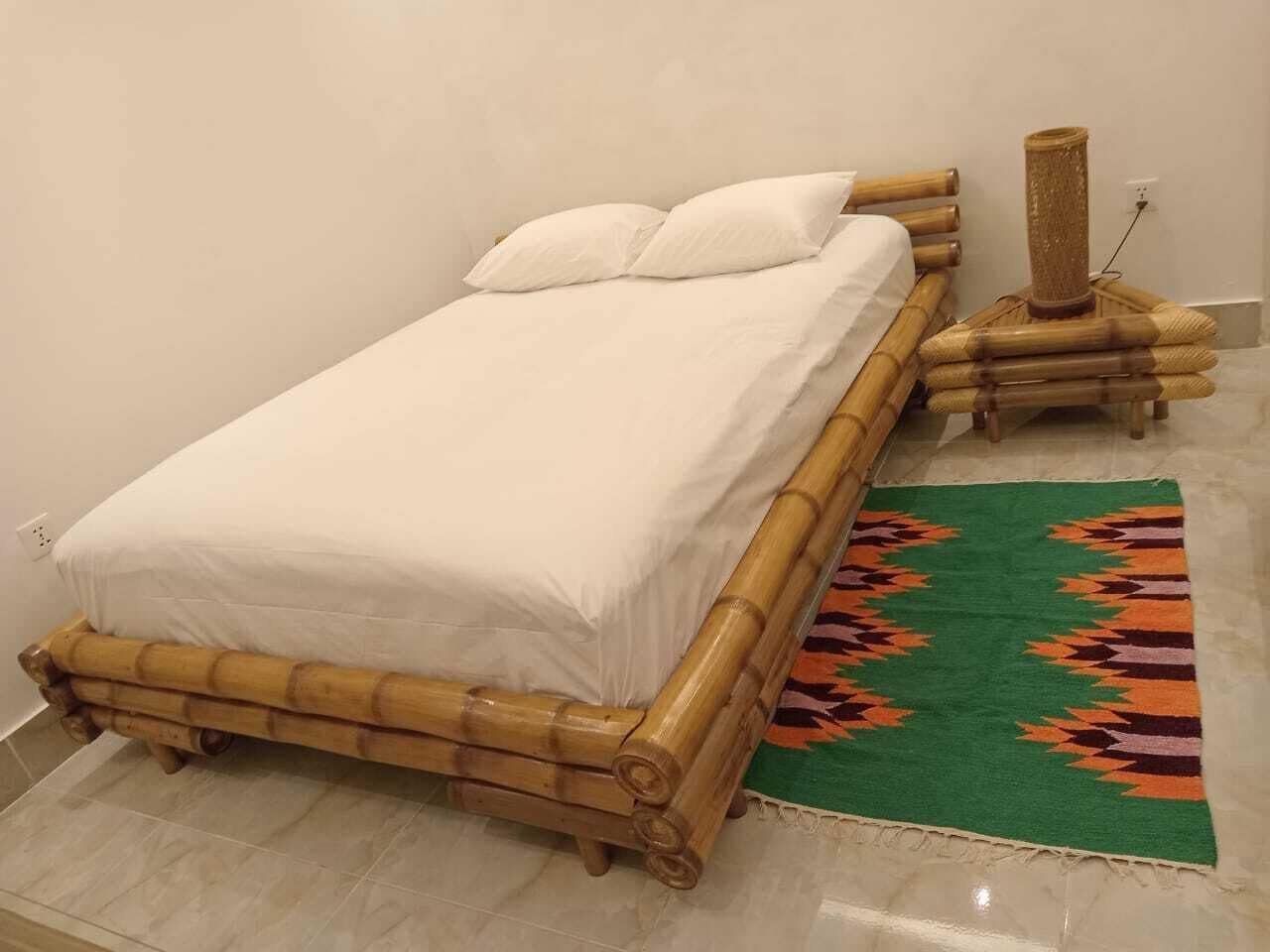 A single bed with a white mattress and pillows, a bamboo bed frame, and a green patterned rug.