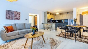 A modern living room with a sofa, coffee table, and a dining area with chairs and a table. A kitchen with a marble countertop and wooden cabinets is visible in the background. - Spacious Loft in Lavington With Pool + Gym & Playground (Nairobi)