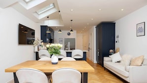 A modern kitchen with a dining area, featuring a wooden table, white chairs, and a sofa.