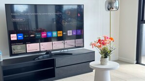 Ein modernes Wohnzimmer mit einem Flachbildfernseher, der ein Heim-Dashboard anzeigt, einem kleinen Tisch mit Blumen und braunen Ledersofas.